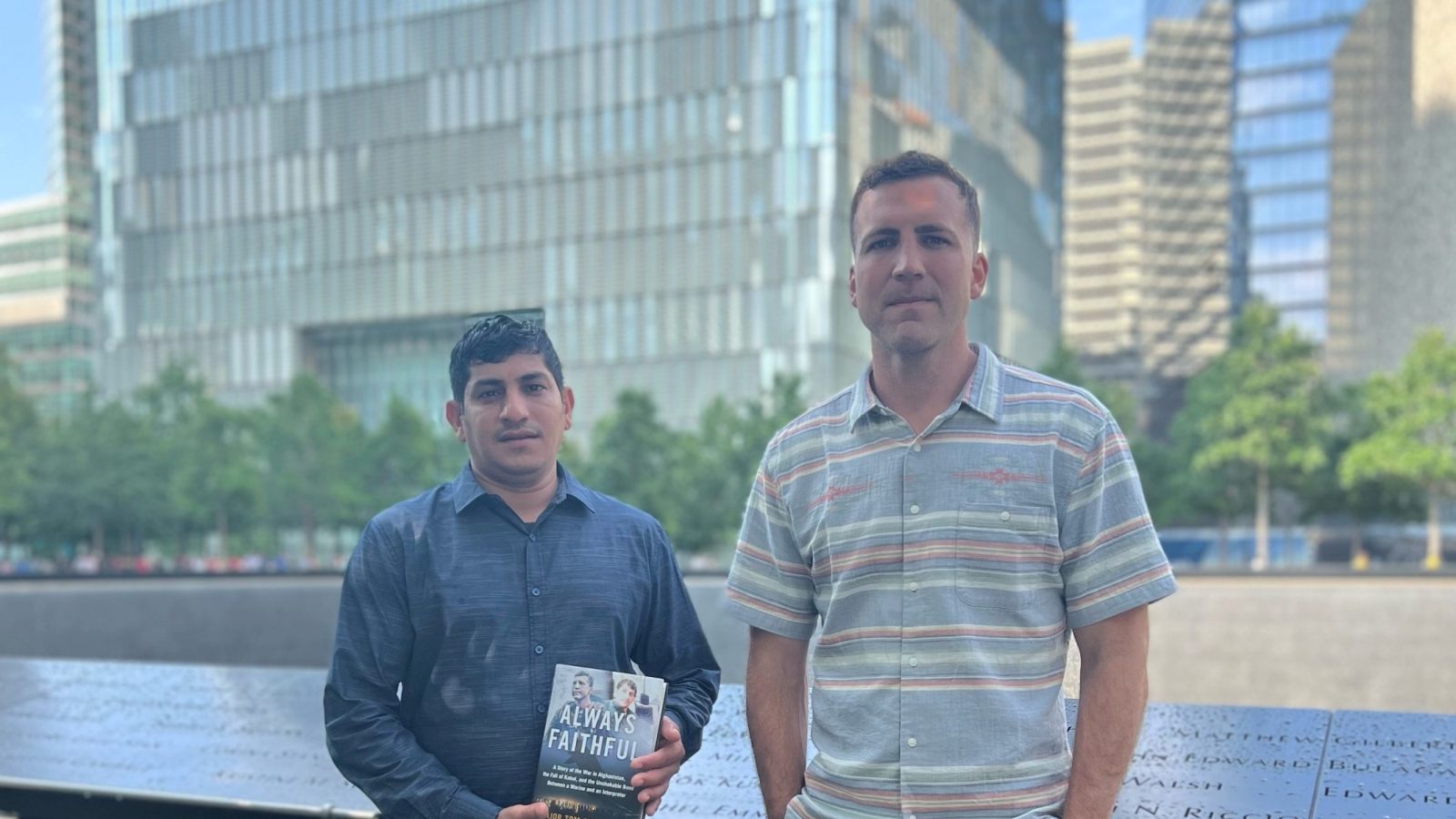 Two men stand next to each other in front of blue river and skyscrapers. The man on the left holds a book and is wearing a blue long-sleeved shirt and khakis. The man on the right wears a short-sleeve button-down and jeans.