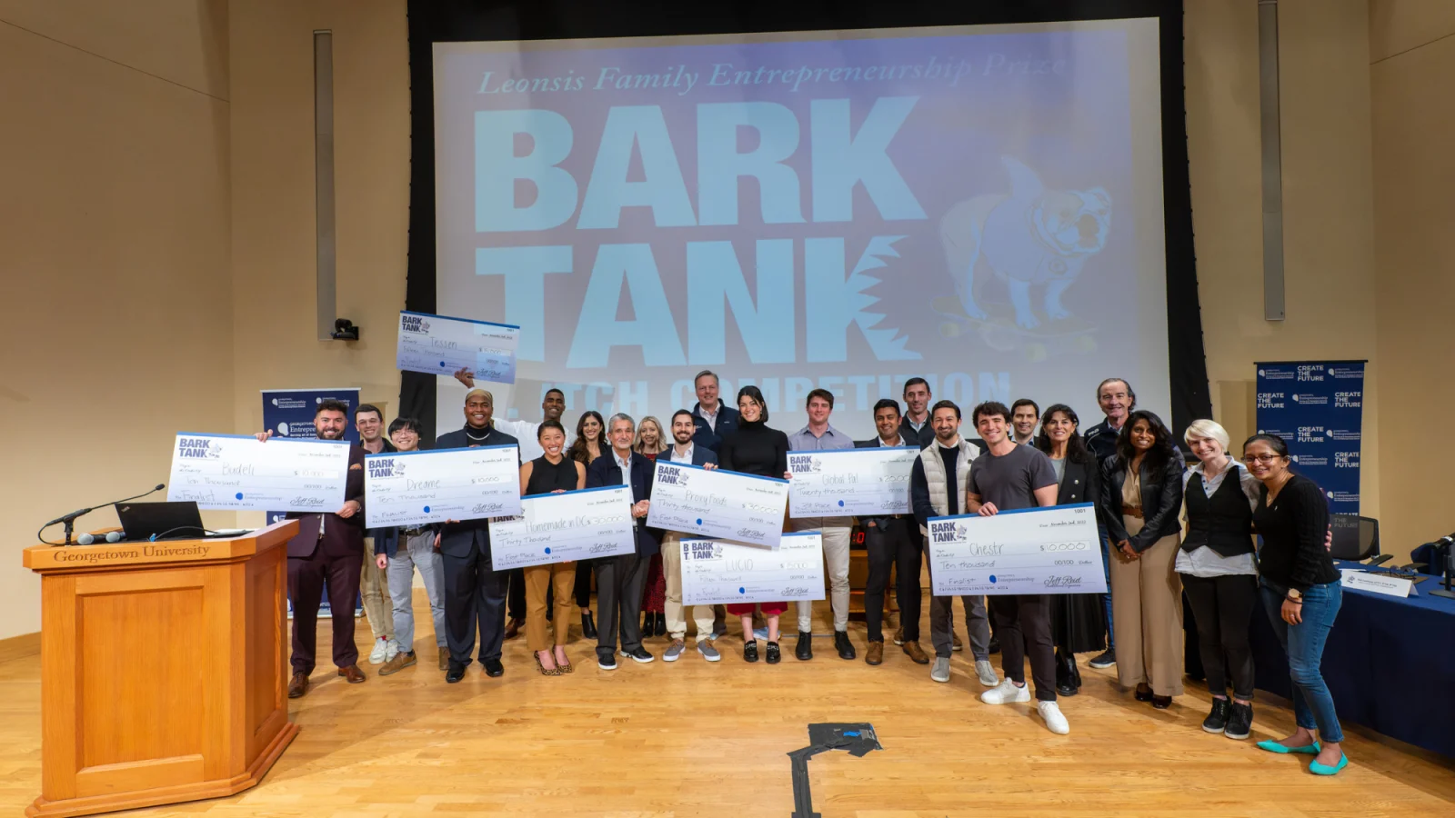 A group of students stand on stage holding giant checks with a screen behind them that says 