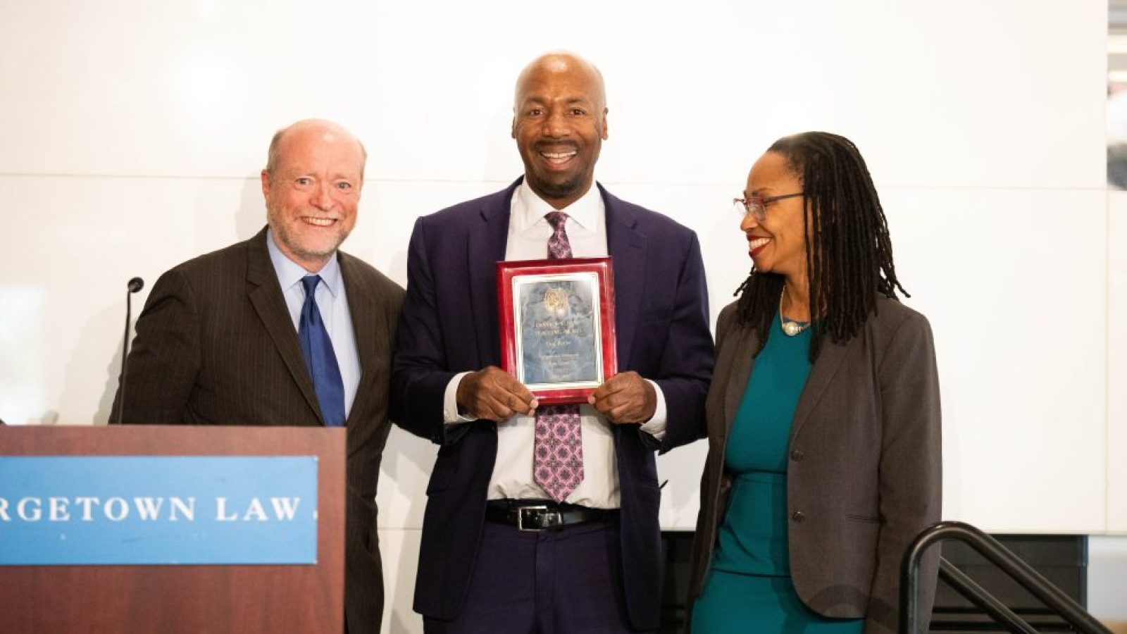 Three members of Georgetown Law stand beside a podium. The professor in the middle holds an award and smiles, wearing a black suit with a tie. The professor on the right wears a blue dress and a blazer, and the Law School Dean on the left wears a black suit and a blue tie.