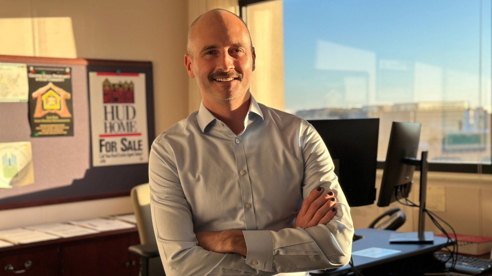 Brian McCabe (SFS&#039;02) stands in his office wearing a blue button-down. Behind him to his right is a window with blue skies, and behind him to his left are posters that say &quot;HUD&quot; on them.