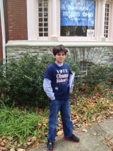 An image of Alex Grass (C'24) at age 7 wearing a blue shirt over a long-sleeved shirt with jeans. His short says "Vote Obama Biden."