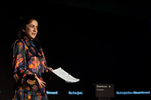 Caitlin Cassidy, a Georgetown alumna, stands on a black stage performing for the New York Times' Climate Forward program. She wears an orange and black flowered dress and holds a script in her left hand.