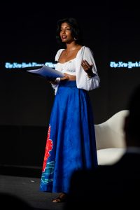 Ashanee Kottage (SFS'22) performs on a black stage at the New York Times Climate Forward program. She wears a vibrant blue skirt with a pink flower on the side and a white blousy shirt. She holds a script in her hands.
