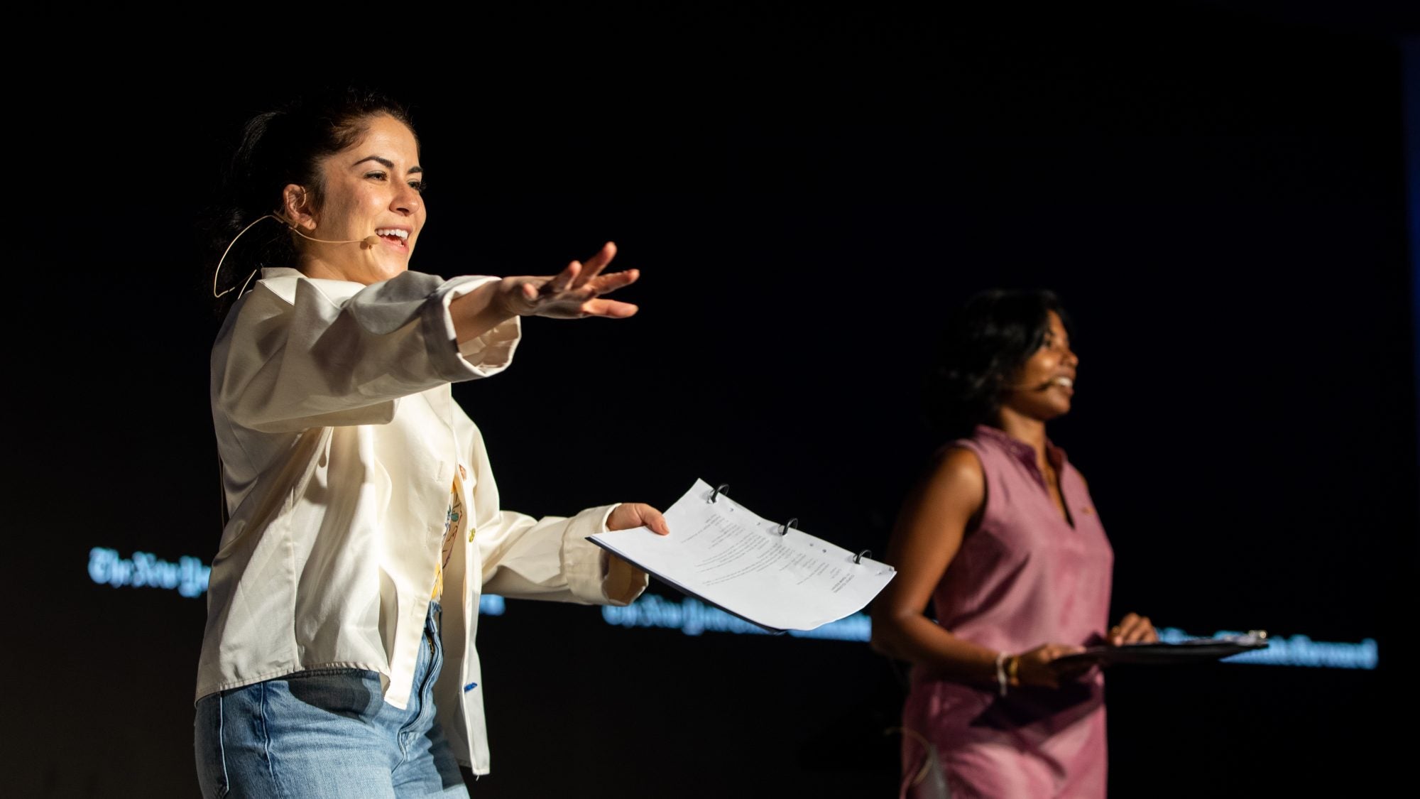 Two women stand on a stage with a black background at the New York Times&#039; Climate Forward program. One woman wears a white jacket and blue jeans, and other other woman, on the right, wears a pink dress.