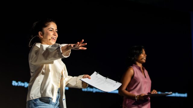 Two women stand on a stage with a black background at the New York Times' Climate Forward program. One woman wears a white jacket and blue jeans, and other other woman, on the right, wears a pink dress.