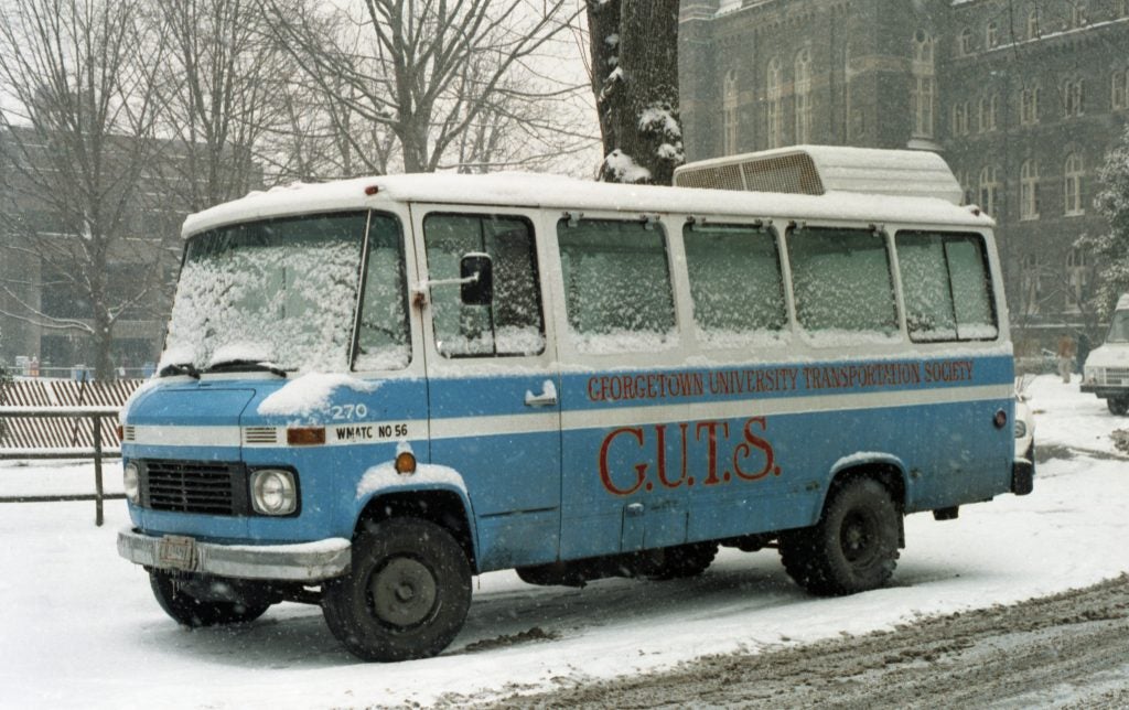 A 1984 photograph of a light blue and white Georgetown GUTS bus covered in snow. The GUYS bus is parked in Healy Circle, and the road is covered in snow.