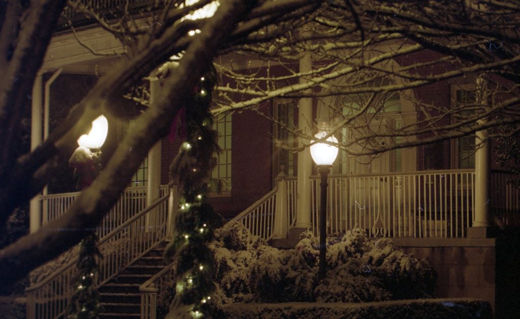 The steps of Old North as seen through tree branches covered in snow. Two lamp posts light up the quad, and the lamp posts have white Christmas lights wrapped around them.
