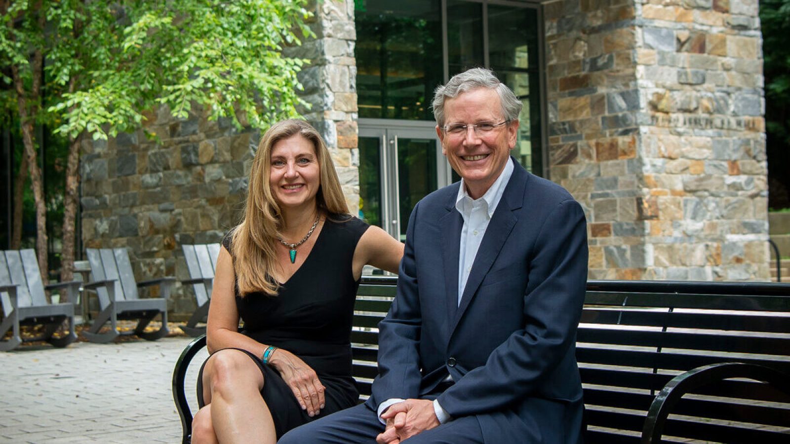 Sheila McMullan (left) wears a black dress and John Monahan (right) wears a suit as they sit on a bench in front of a stone building