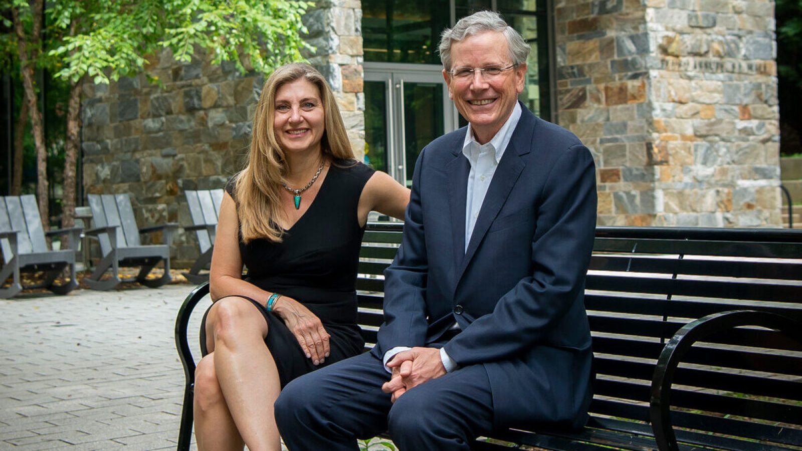 Sheila McMullan (left) wears a black dress and John Monahan (right) wears a suit as they sit on a bench in front of a stone building