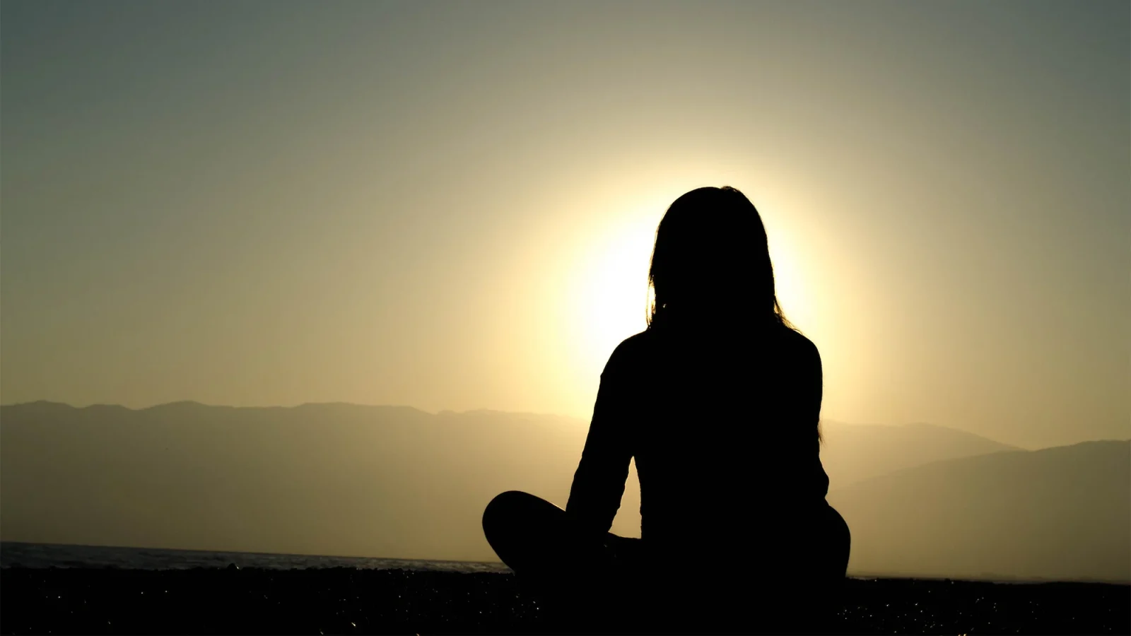 Woman sits cross-legged in shadow facing the sun on the horizon
