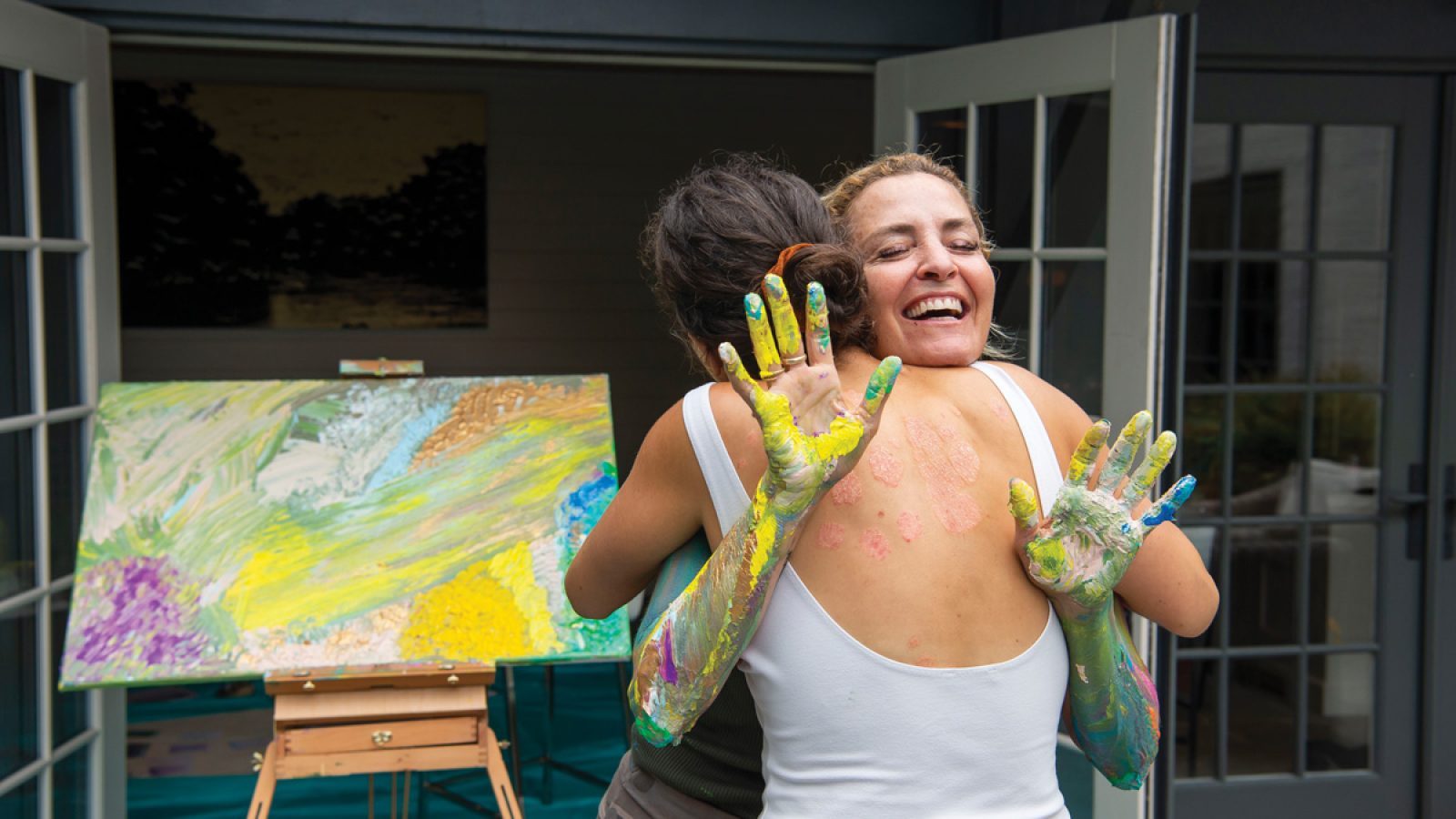 Two women hug each other in front of a painted easel. One of the women facing the camera holds out her painted hands.
