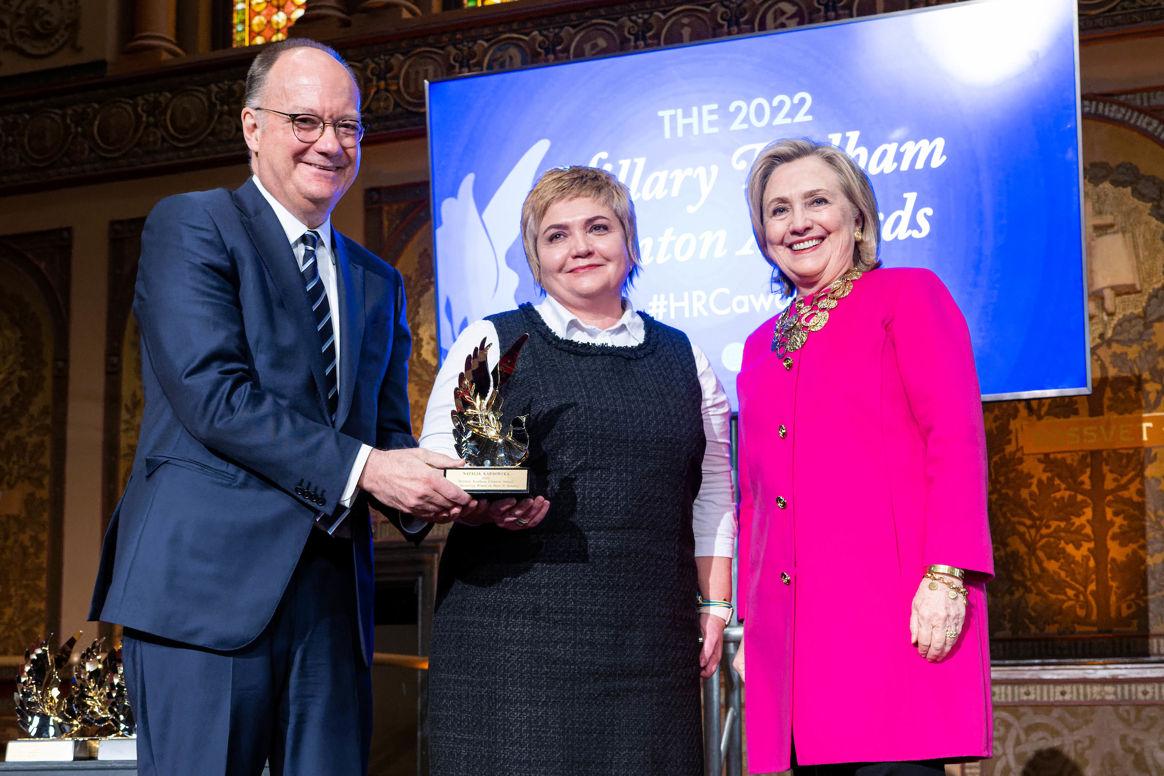 Jack DeGioia, Natalia Karbowska and Hillary Clinton hold an award onstage. 