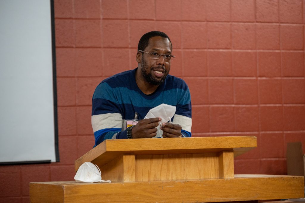 Colie “Shaka” Long speaks at a podium at the Prison Scholars Program’s end-of-semester celebration at the DC Jail in December 2022. He is wearing a blue-and-white-striped long-sleeve shirt and glasses.
