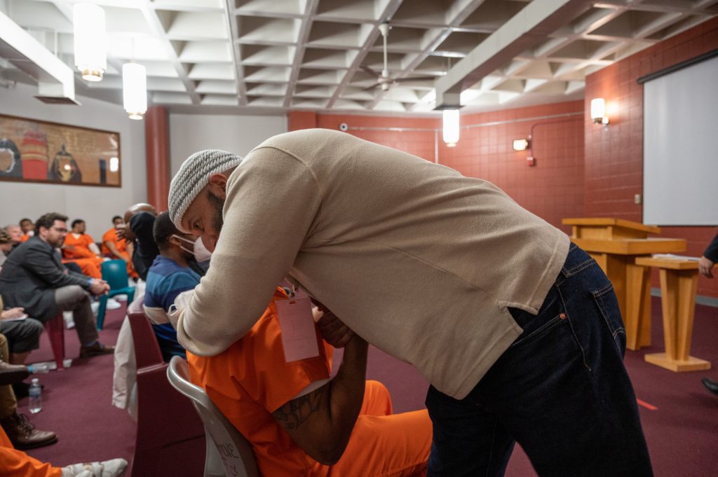 Adnan Syed bends down to hug a scholar in Georgetown's Prison Scholars Program during the program's end-of-the-semester celebration at the DC jail. Behind him, scholars wear orange jumpsuits. 