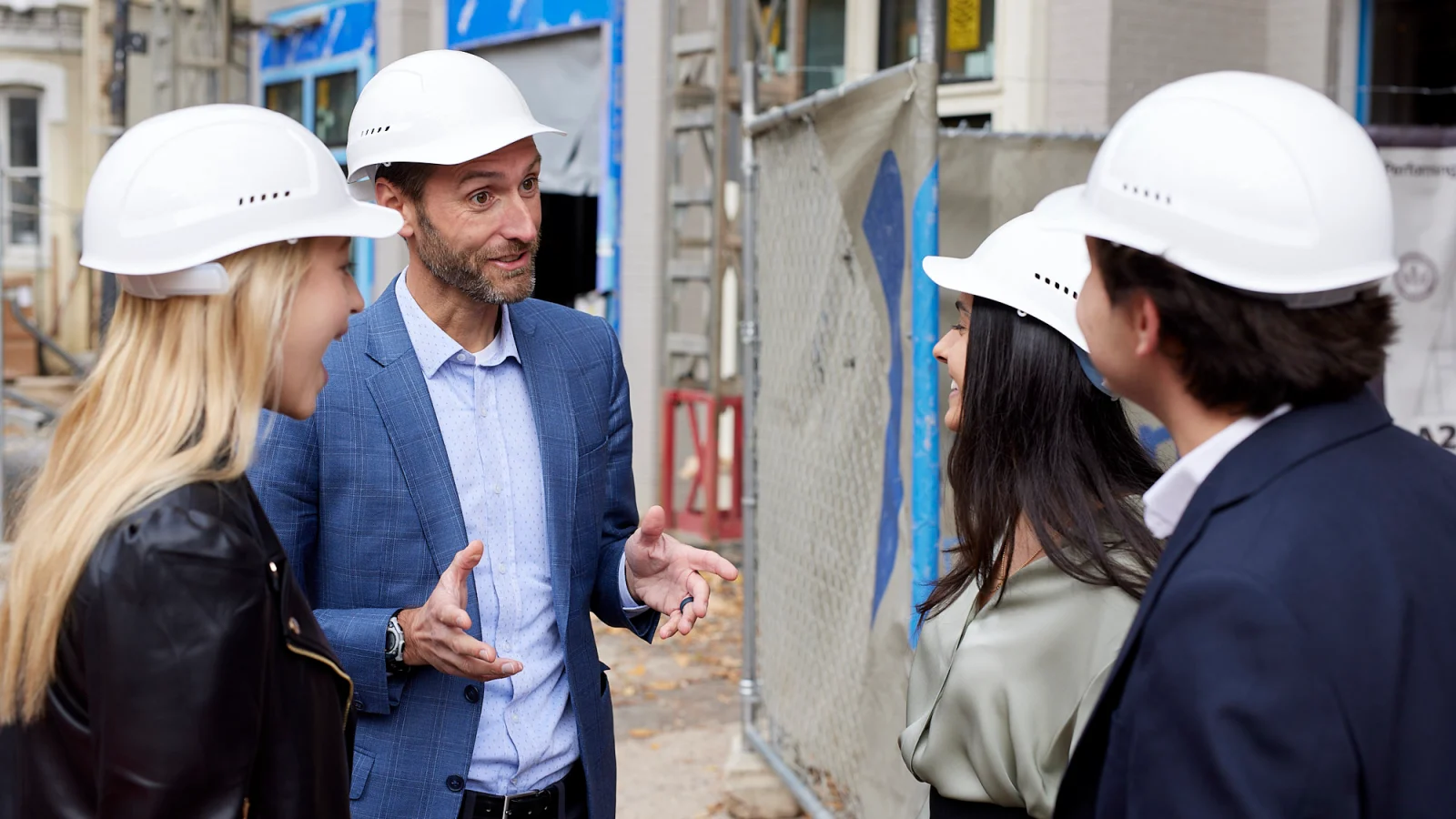 Two men and two women stand in a circle talking. They are dressed in formal business wear and all are wearing white hard hats. Behind them is a construction zone.