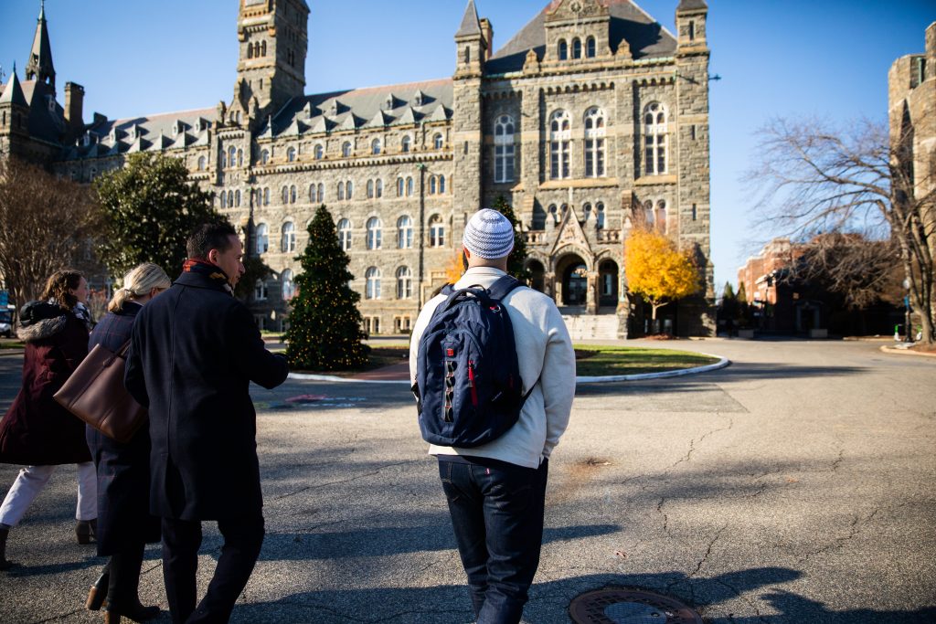 Adnan Syed (right) walks toward Healy Hall, a Gothic-style building on Georgetown's main campus, with three others. He is wearing a cream-colored sweater and a backpack. In front of him is a Christmas tree and a bright blue sky.