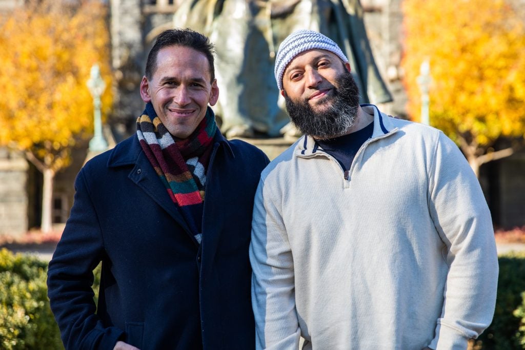 Adnan Syed (right) poses for a picture with Marc Howard (left), director of the Prisons and Justice Initiative. Syed is wearing a cream-colored sweater and a blue-and-white-striped kufi, or Muslim prayer hat. Howard is wearing a blue suit and a striped scarf.