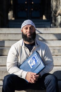 Adnan Syed, a program associate in the Prisons and Justice Initiative, sits on the front steps of Healy Hall. He is wearing a cream-colored zip-up sweater, a blue-and-white-striped kufi, or Muslim prayer hat, and holding a blue binder with a Georgetown bulldog sticker on it. 