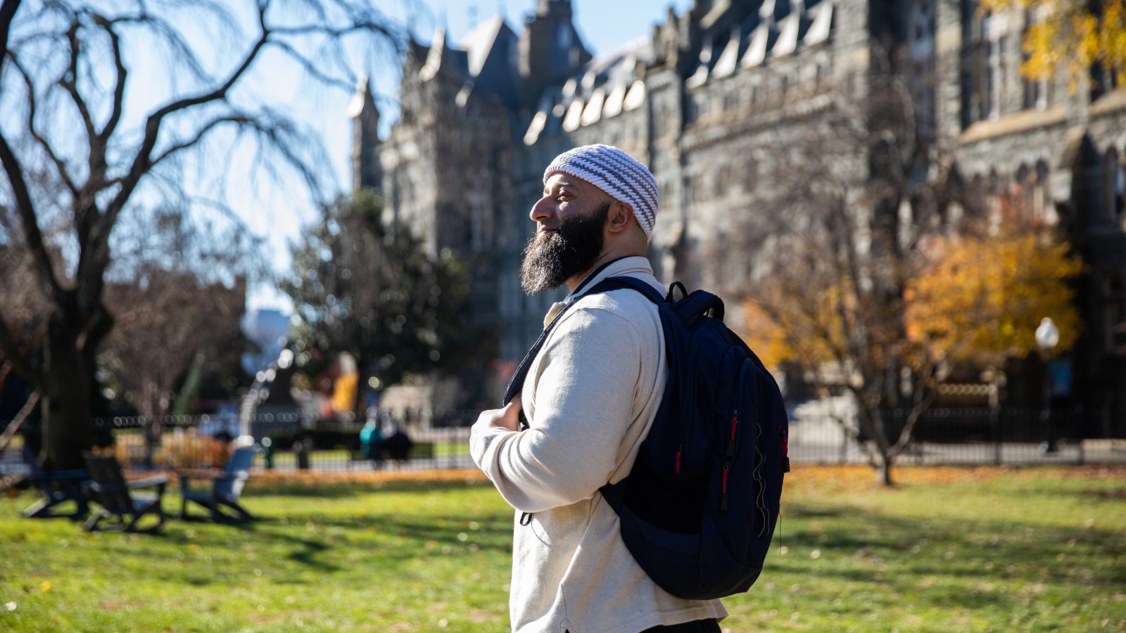 Adnan Syed, a program associate in the Prisons and Justice Initiative, wears a cream-colored sweater, a blue-and-white-striped kufi, or Muslim prayer hat, and a black backpack. He stands on the front lawn of Georgetown with Healy Hall, a Gothic-style building behind him and looks forward.