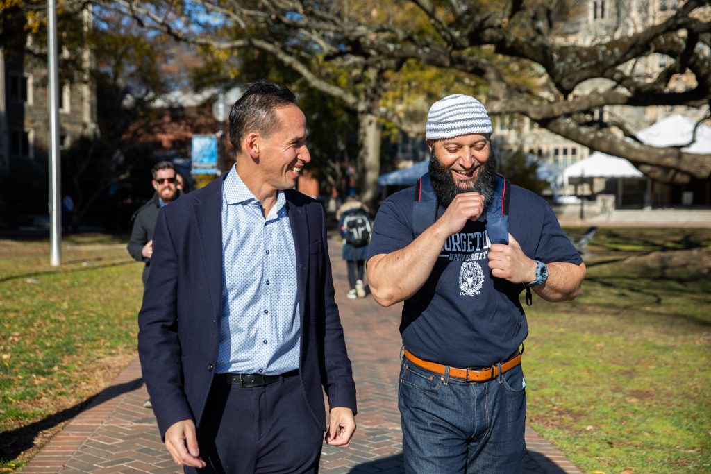 Adnan Syed (right) laughs while walking next to Marc Howard (left), director of the Prisons and Justice Initiative, while walking on a sidewalk on Georgetown's main campus. It's a sunny day and the grass is green. Marc wears a black suit with no tie and Adnan wears a blue Georgetown T-shirt and blue jeans with a Muslim prayer cap.
