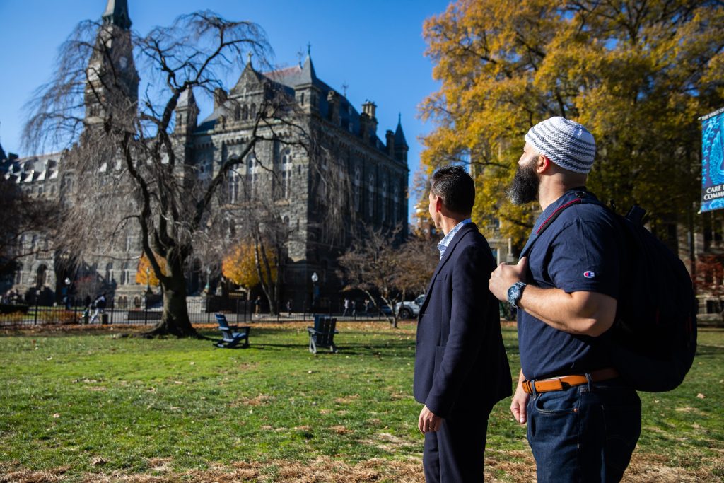 Adnan Syed (right) stares up at Healy Hall, a Gothic-style building on Georgetown's main campus, next to Marc Howard (left), director of the Prisons and Justice Initiative. The sky is bright blue behind Healy Hall. Marc wears a black suit with no tie and Adnan wears a blue Georgetown T-shirt and blue jeans with a Muslim prayer cap.