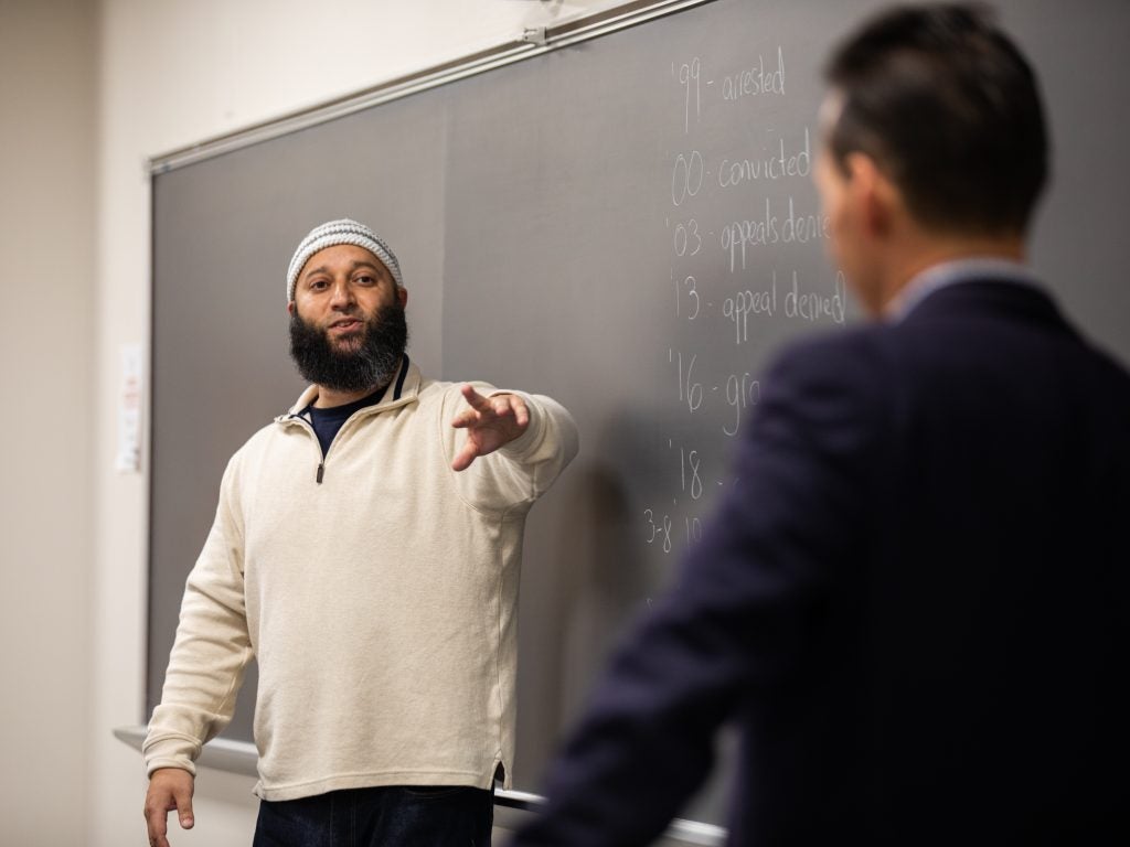 Adnan Syed (left) stands in front of a chalkboard with his left arm outstretched. He is wearing a cream-colored sweater and a blue-and-white-striped kufi, or Muslim prayer hat. In the foreground with his back to the camera is Marc Howard, director of the Prisons and Justice Initiative, who is wearing a blue suit.