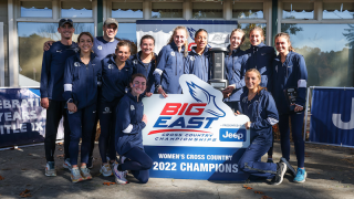 Women&#039;s cross country team in dark blue sweat suits stand behind a Big East 2022 Champions sign