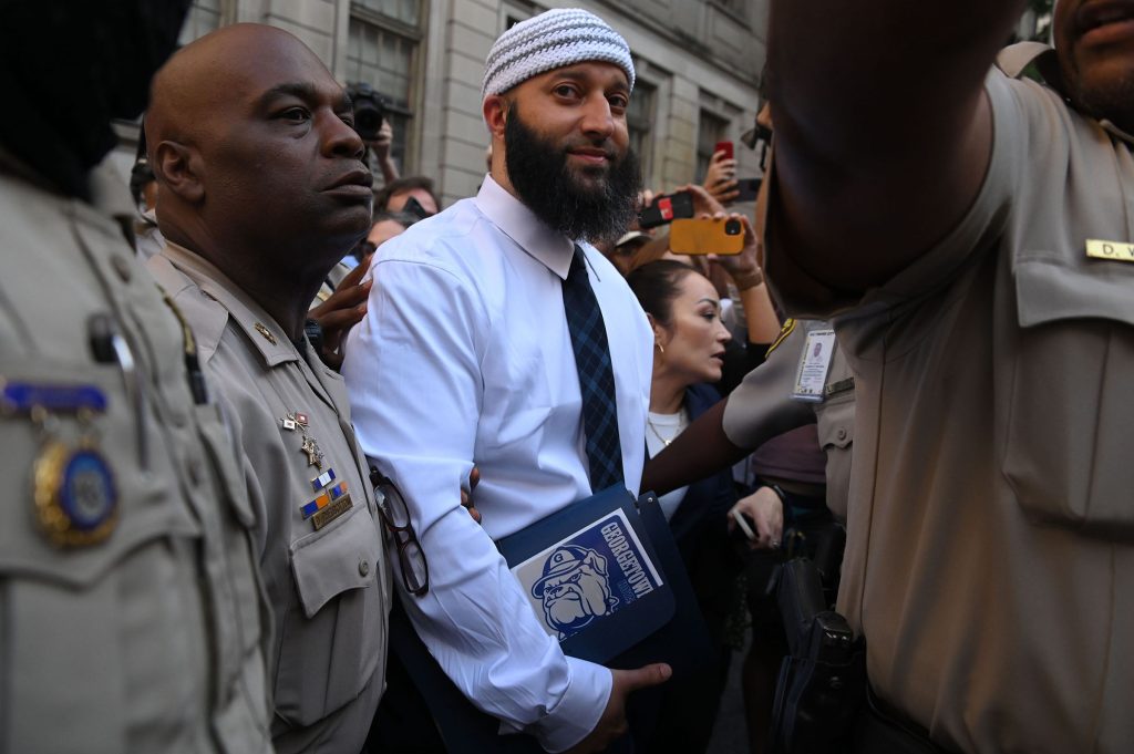 Adnan Syed leaves the courthouse after being released from prison Monday, Sept. 19, 2022, in Baltimore. He is wearing a white button-down shit and a tie, and holding a blue binder with a Georgetown bulldog sticker on it.