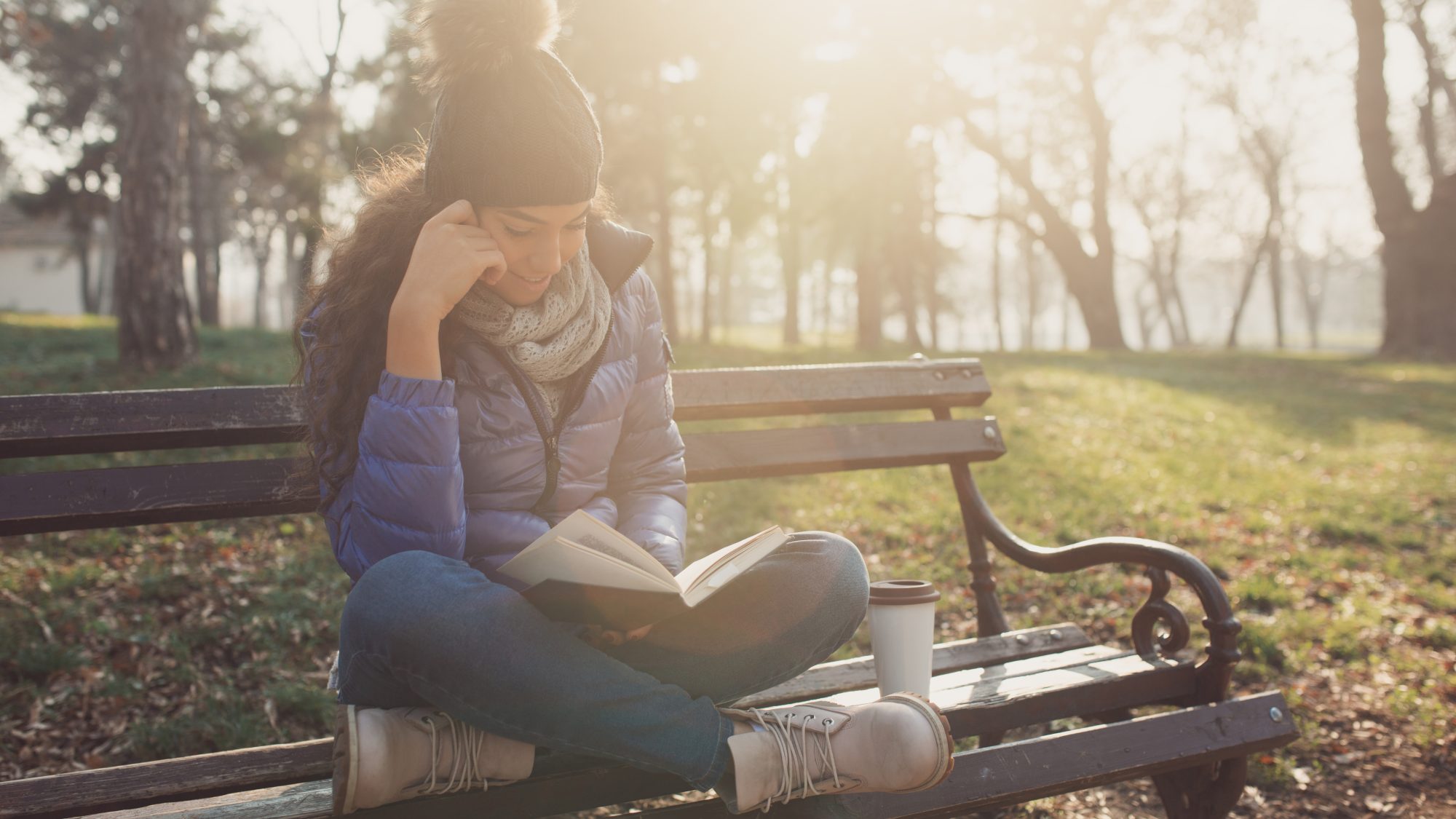 A young woman sits on a park bench cross-legged reading a book. She is wearing a pompom hat, a purple winter jacket and jeans. Next to her on the bench is a coffee cup.