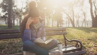 A young woman sits on a park bench cross-legged reading a book. She is wearing a pompom hat, a purple winter jacket and jeans. Next to her on the bench is a coffee cup.