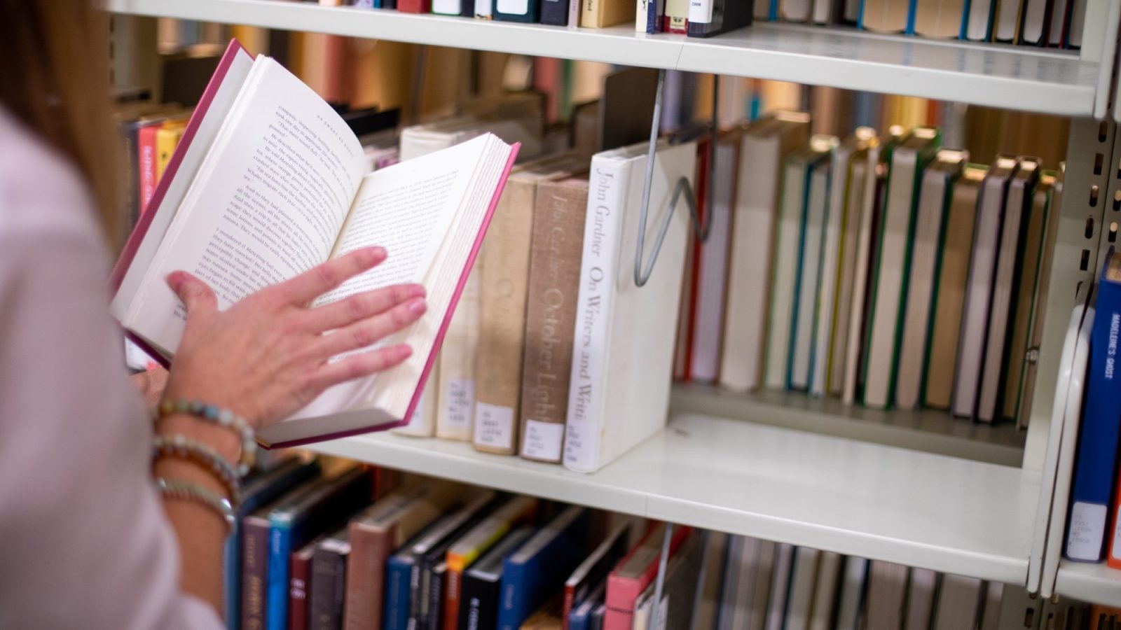 A woman rests her hand between pages of an open book. Behind her is a library shelf of books. The woman is wearing a purple shirt and two bracelets.