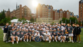 Men&#039;s Lacrosse team celebrates in a group photo with the sun setting behind the field