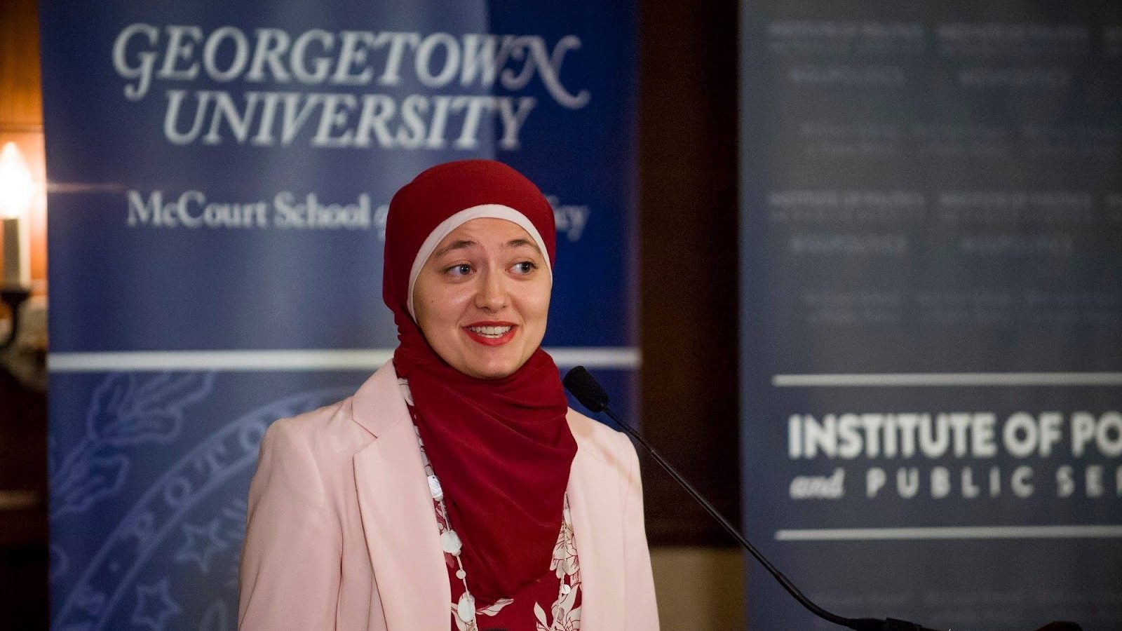 Ruwa Romman, a graduate of the McCourt School, wears a red hijab and a pink suit while speaking at a podium in front of a blue sign that says &quot;Georgetown University.&quot;