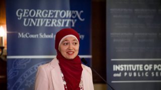 Ruwa Romman, a graduate of the McCourt School, wears a red hijab and a pink suit while speaking at a podium in front of a blue sign that says &quot;Georgetown University.&quot;