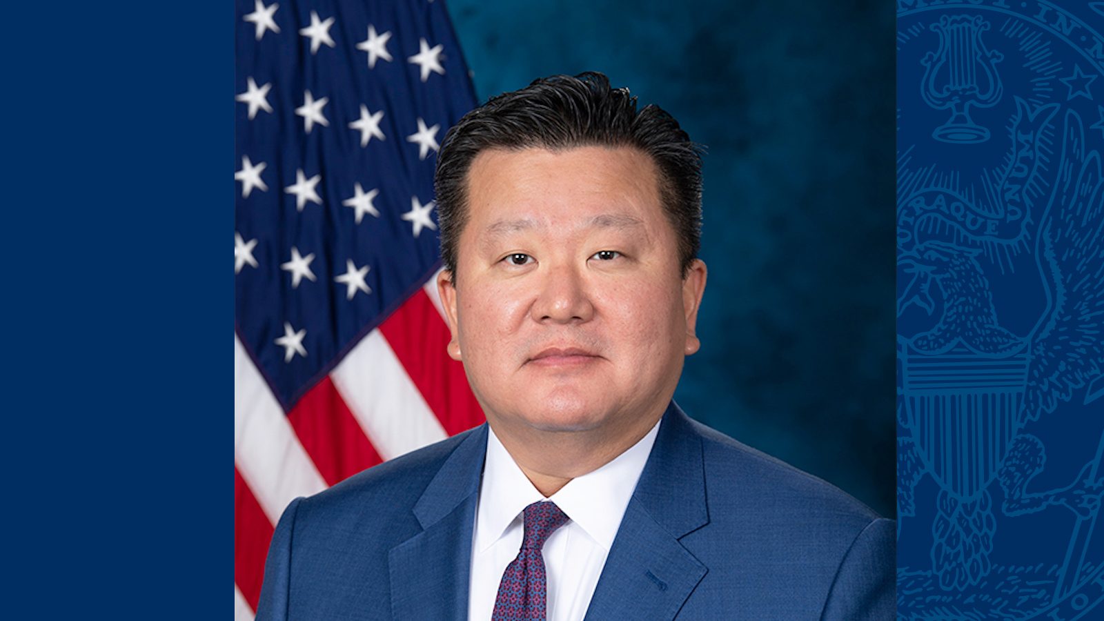 Ryung Suh wears a blue suit and purple tie in front of an American flag