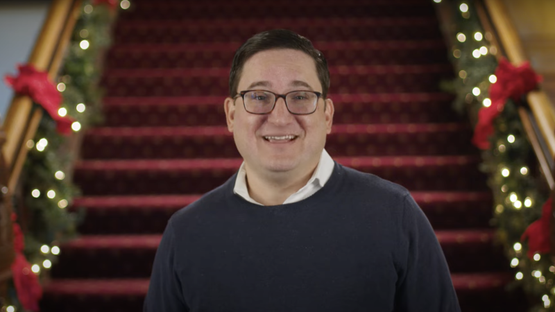 Jonah Perlin wears a sweater and glasses in front of red carpeted stairs with garland, lights and red ribbon on the railings.
