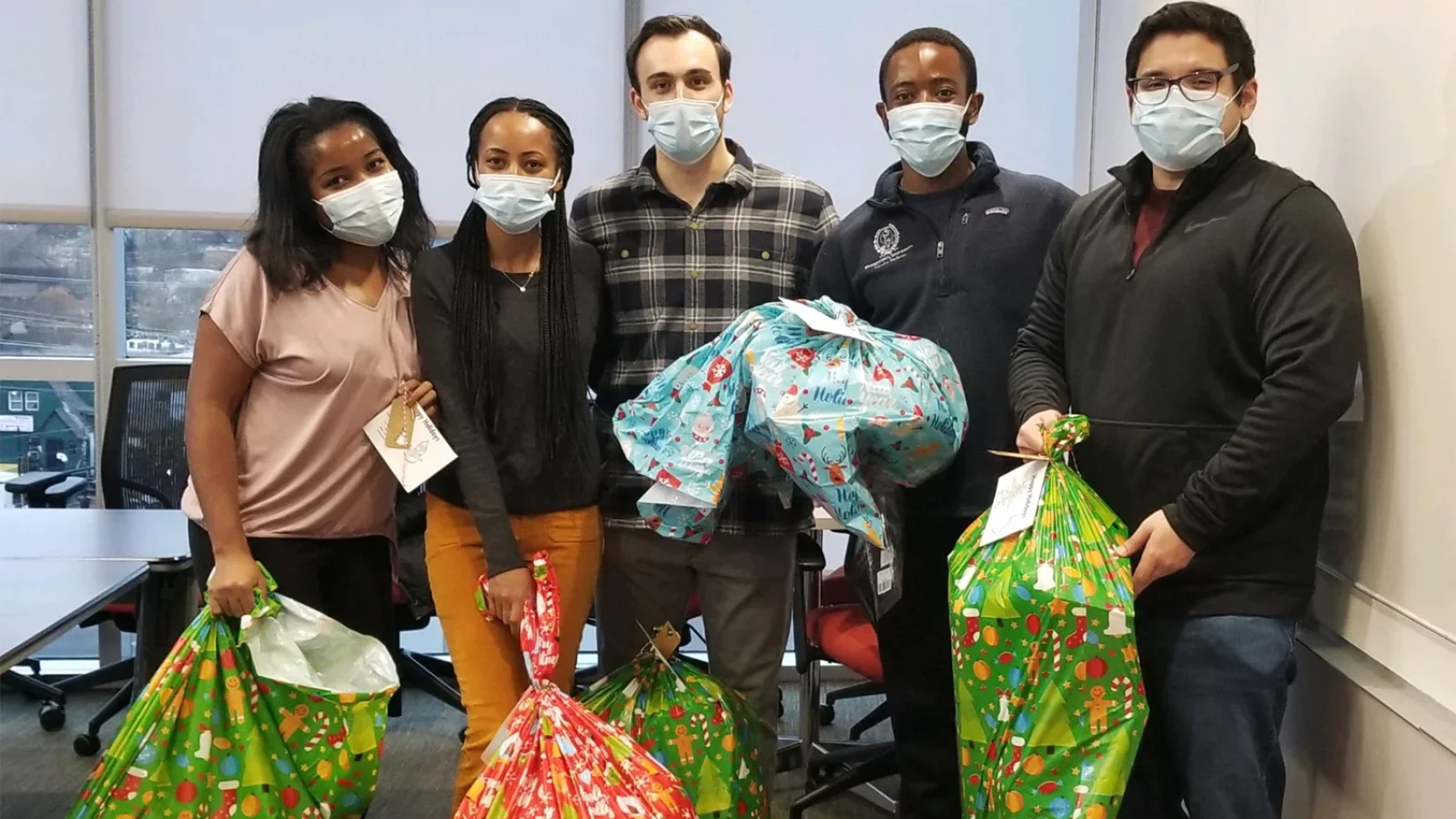 Five students wear masks and hold wrapped presents inside a conference room.