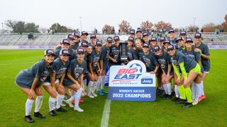 Women&#039;s Soccer team celebrates in a group photo on the field behind a sign that says &quot;2022 Champions&quot;