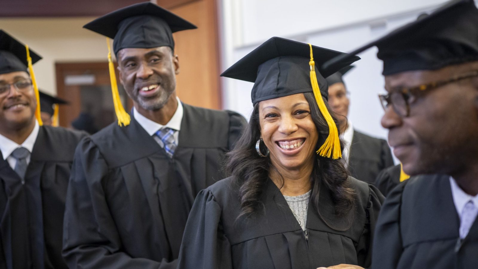 A group of returning citizens wear black graduation robes and caps with yellow tassels dangling on one side. They are smiling during an event celebrating their graduation from Georgetown&#039;s Paralegal Program.