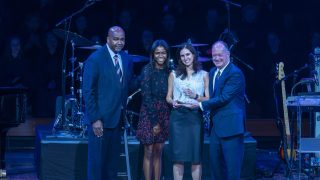 Four people stand on stage and smile toward the audience. President DeGioia, standing on the far right, is handing an award to the woman standing next to him.
