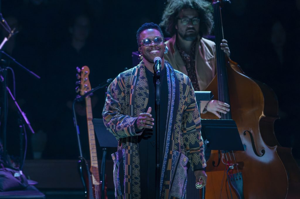 Leslie Odom, Jr., performs on stage during his performance at the Let Freedom Ring Celebration. Behind him is his band; one man is holding a bass. Leslie wears sunglasses and a long tan patterned coat over black pants and a black shirt.