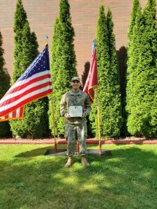 On a sunny day, Arthur Milhomens (B’24) wears a military uniform and stands in front of four trees and two American flags. He holds an award in his hand.