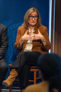 Rachel Bronson, the CEO of the Bulletin of Atomic Scientists, the nonprofit organization that oversees the Doomsday Clock, sits on a chair during a panel discussion at Georgetown. She rests one arm on her right leg and holds the other up as she talks. She is wearing a tan jacket, navy trousers, and has her glasses pulled up in her hair.