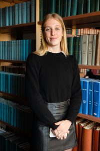 Ellie Cotter (G’23), a graduate student in Georgetown’s Conflict Resolution program, poses for a picture behind bookshelves at the Jan. 24 event on campus. She has long blonde hair and wears a black sweater and a leather skirt.