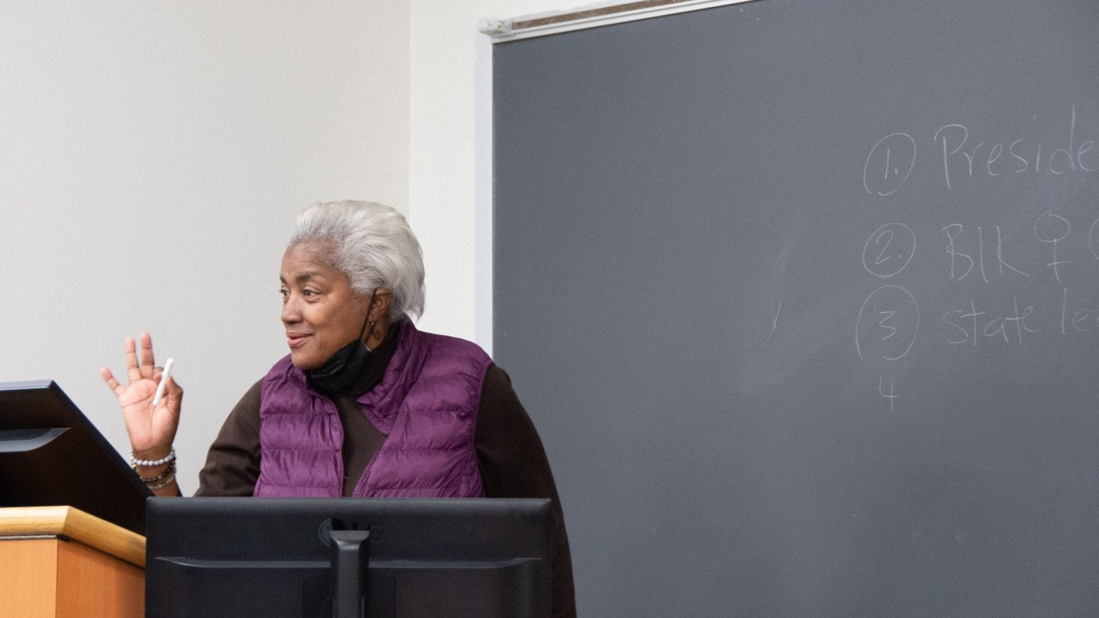 Donna Brazile, an adjunct professor at Georgetown, stands behind a podium and a blackboard while teaching a class. She is wearing a brown sweater with a purple vest over it.