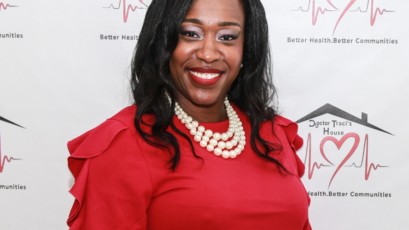 Dr. Traci Thompson (C&#039;96) stands in front of a white background with her clinic&#039;s logo &quot;Dr. Traci&#039;s House&quot; written behind her. She has long black hair and wears pearls, a red long-sleeve shirt, and a black skirt. Her nails are painted pink.