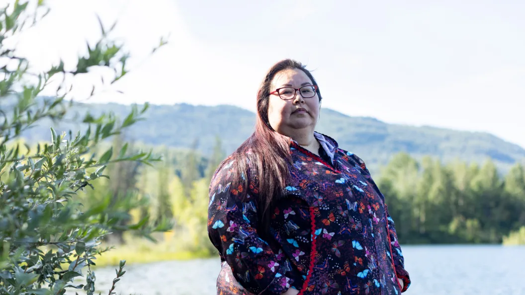 Angel Dotomain (B&#039;99) wears a purple button-up shirt with blue, red, and multi-colored butterflies on it and rectangle glasses. She stands in front of a green mountain, which is blurred behind her.
