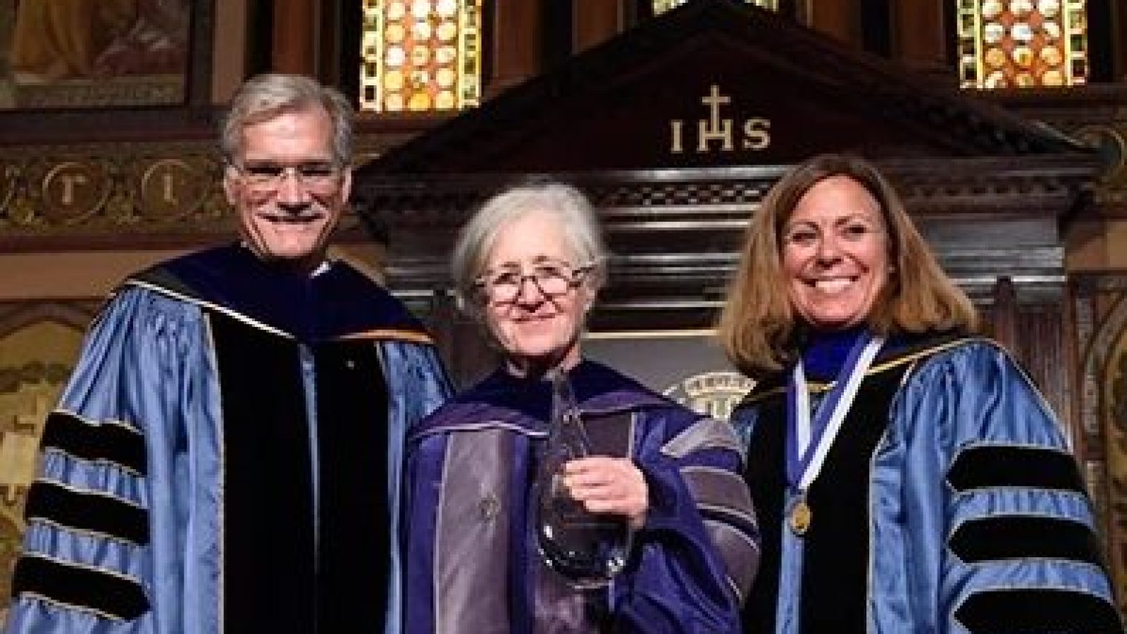 Professor Maxine Weinstein (center) wears a purple graduation robe at a commencement. She stands on the stage of Gaston Hall next to Provost Robert Groves, who wears a blue graduation robe with three black stripes on the sleeves, and another woman wearing a blue graduation robe with three black stripes on the sleeves.