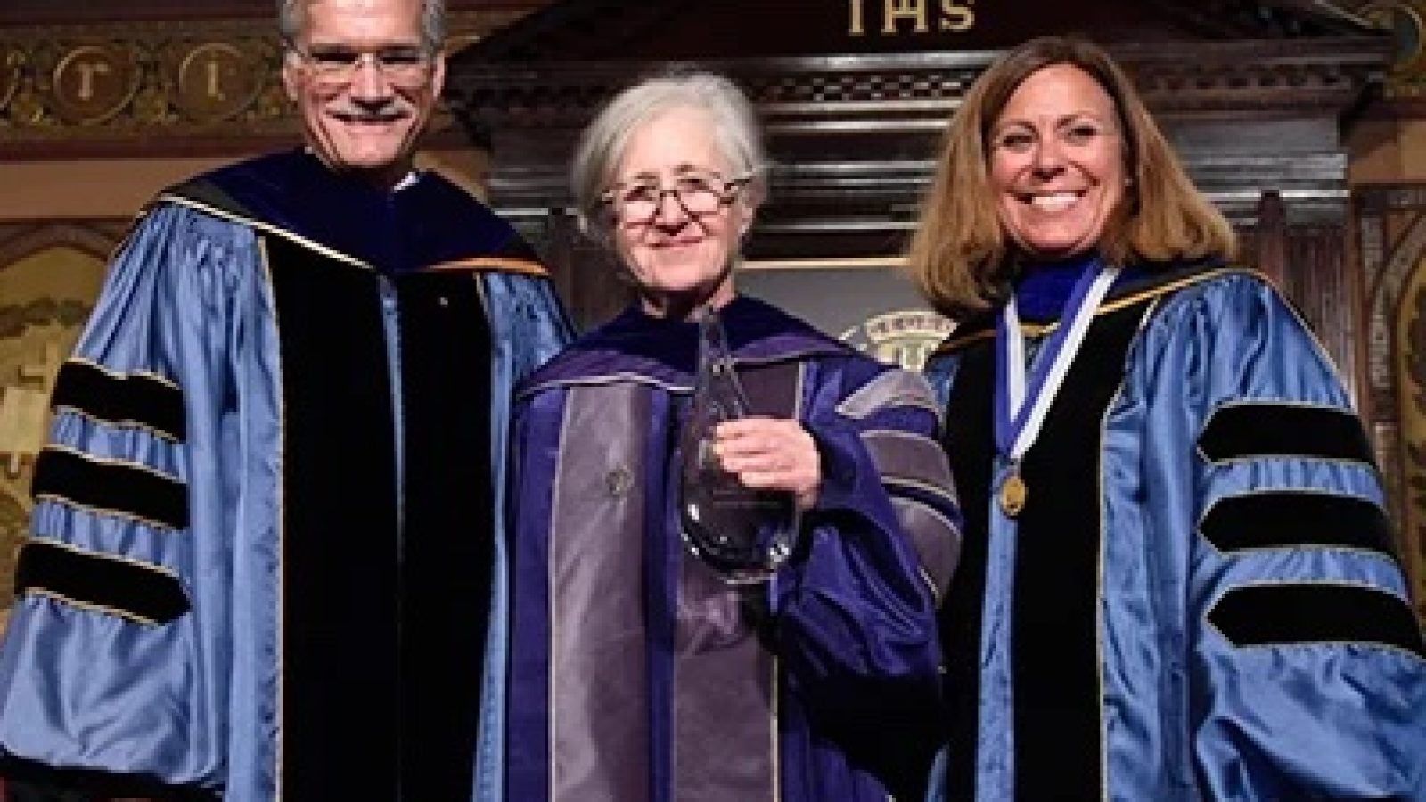 Professor Maxine Weinstein (center) wears a purple graduation robe at a commencement. She stands on the stage of Gaston Hall next to Provost Robert Groves, who wears a blue graduation robe with three black stripes on the sleeves, and another woman wearing a blue graduation robe with three black stripes on the sleeves.