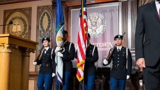 Students wearing military regalia stand on stage in Gaston Hall, holding an American flag.